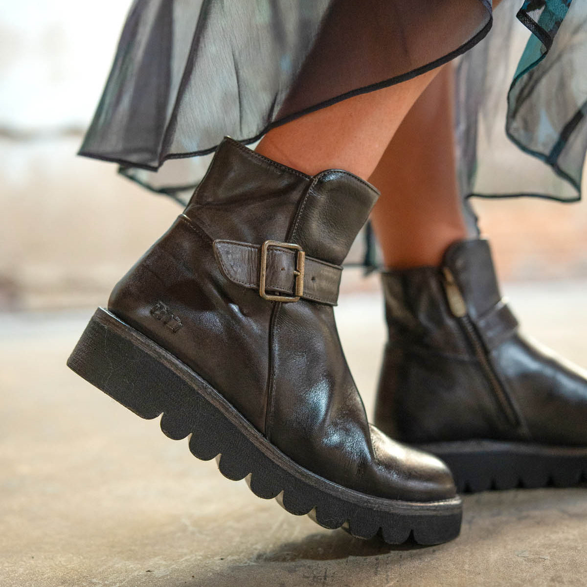 Close-up of a person wearing Bed Stu Nudge II black leather ankle boots with a side buckle, adjustable strap, and thick ridged soles, styled with a sheer, flowy skirt.