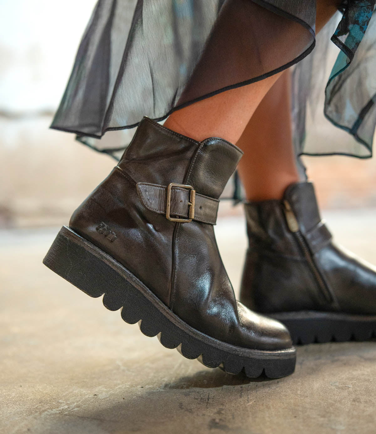 Close-up of a person wearing Bed Stu Nudge II black leather ankle boots with a side buckle, adjustable strap, and thick ridged soles, styled with a sheer, flowy skirt.