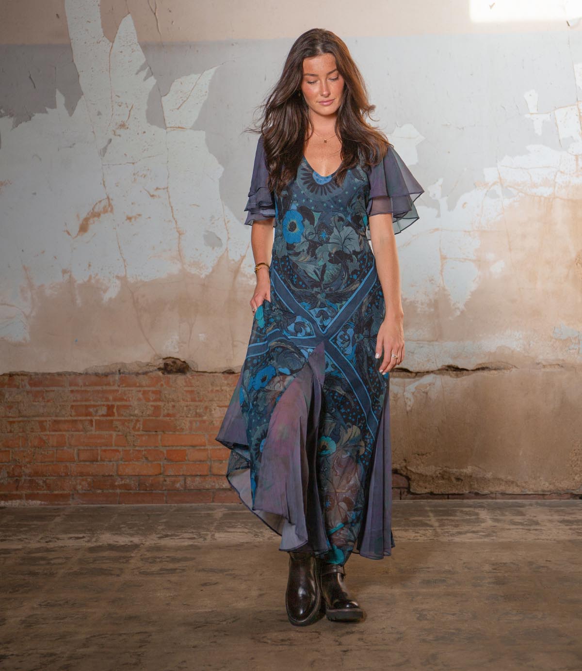 A woman with long brown hair wears Bed Stu’s Nudge II black ankle boots and a blue floral dress as she stands on a concrete floor before a distressed wall.