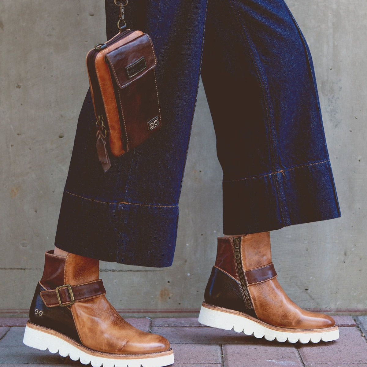 A person wearing the Nudge II brown leather ankle boots with white soles by Bed Stu and blue wide-leg jeans, holding a small brown leather shoulder bag with an adjustable strap, walks on a brick sidewalk.
