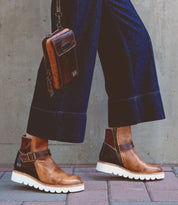 A person wearing the Nudge II brown leather ankle boots with white soles by Bed Stu and blue wide-leg jeans, holding a small brown leather shoulder bag with an adjustable strap, walks on a brick sidewalk.