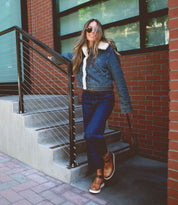 Person in denim jacket and jeans, wearing sunglasses and holding a bag, standing on outdoor steps next to a brick building. They're also sporting the stylish Bed Stu Nudge II ankle boots with an adjustable strap, adding a chic touch to their casual look.