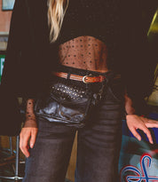 A person wearing the Occurrence black sheer polka dot top, black jeans, and a studded leather belt, with an edgy Bed Stu crossbody bag adorned in sleek studs slung over the waist.