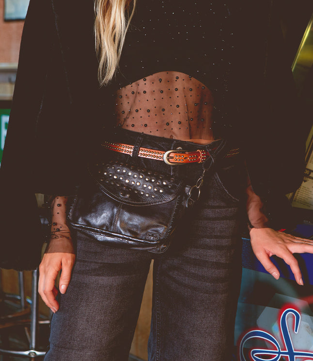 A person wearing the Occurrence black sheer polka dot top, black jeans, and a studded leather belt, with an edgy Bed Stu crossbody bag adorned in sleek studs slung over the waist.