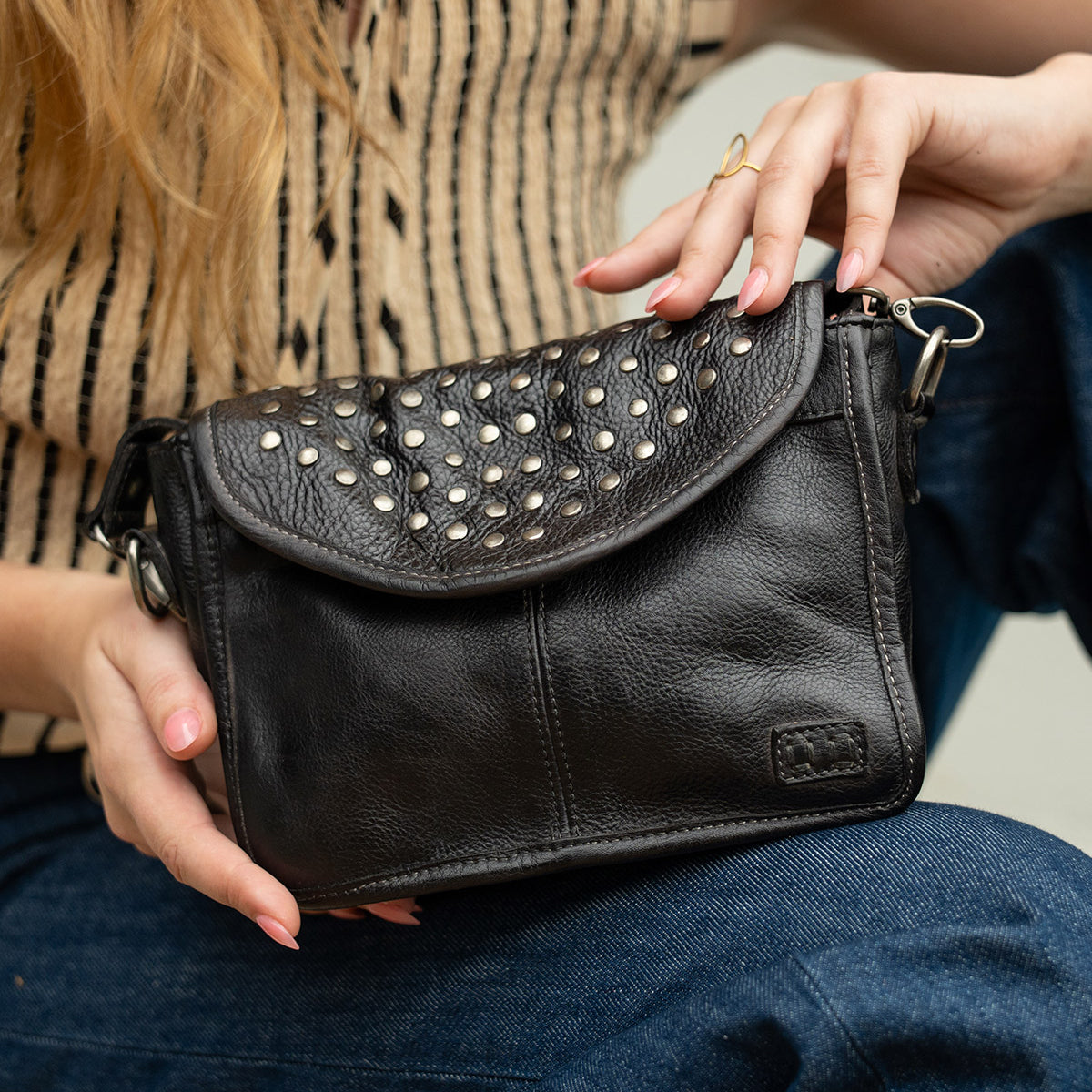 Wearing a striped sleeveless top and jeans, the person holds the Bed|Stü Occurrence—a compact crossbody bag featuring metal stud accents and a flap closure.