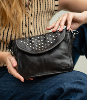 Wearing a striped sleeveless top and jeans, the person holds the Bed|Stü Occurrence—a compact crossbody bag featuring metal stud accents and a flap closure.