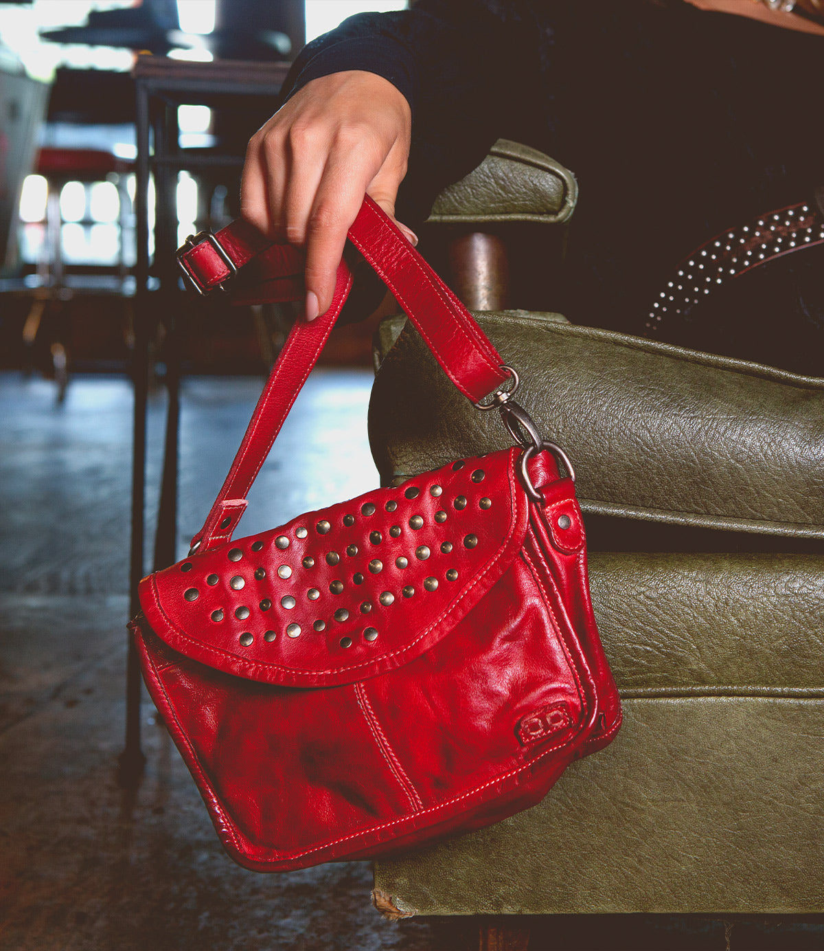 A person sitting on a green chair indoors, showcasing an edgy design with their small red leather Occurrence handbag by Bed Stu adorned with metal studs—a versatile accessory that effortlessly complements any outfit.