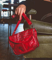 A person sitting on a green chair indoors, showcasing an edgy design with their small red leather Occurrence handbag by Bed Stu adorned with metal studs—a versatile accessory that effortlessly complements any outfit.