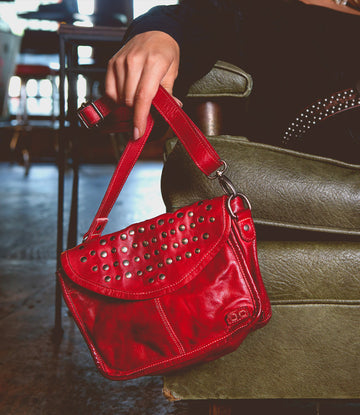 A person sitting on a green chair indoors, showcasing an edgy design with their small red leather Occurrence handbag by Bed Stu adorned with metal studs—a versatile accessory that effortlessly complements any outfit.