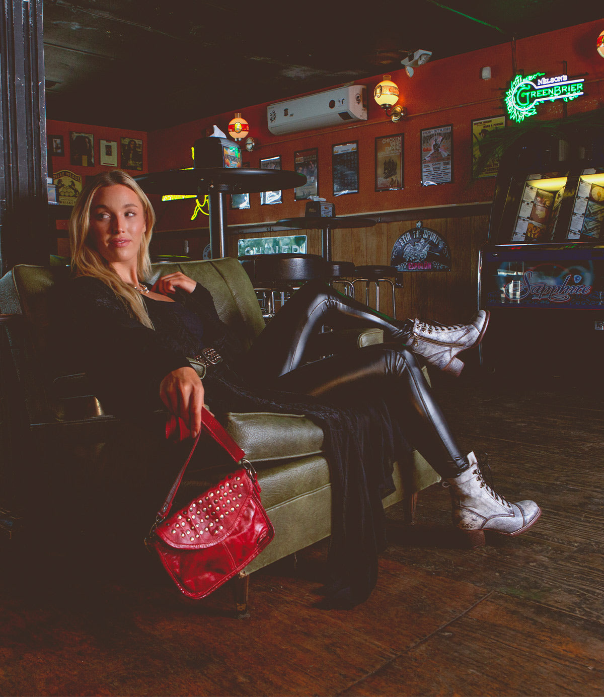A person in leather pants and white boots sits on a green couch in a dimly lit room, holding the edgy Occurrence crossbody purse by Bed Stu. Neon signs and posters decorate the walls.