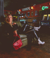 A person in leather pants and white boots sits on a green couch in a dimly lit room, holding the edgy Occurrence crossbody purse by Bed Stu. Neon signs and posters decorate the walls.