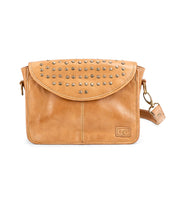 The Bed Stu Occurrence is a tan leather crossbody bag with flap closure, metal stud details, detachable strap, and a small rectangular logo patch on the front—stylish and versatile for any occasion.