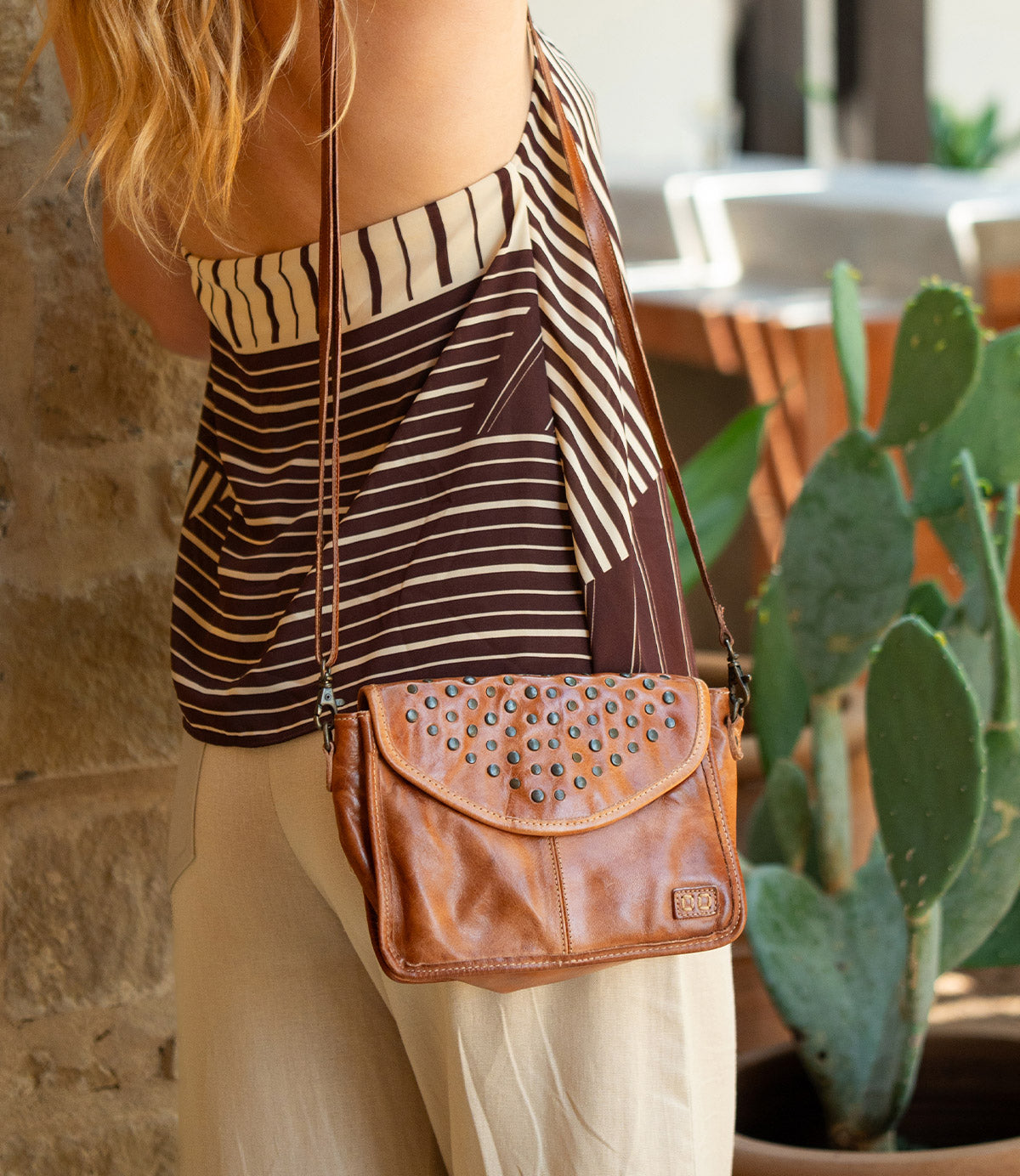 A person wearing a striped top and beige pants stands by a stone wall, carrying the Bed|Stü Occurrence crossbody bag—a compact accessory crafted from brown leather with studded details that perfectly complements the outfit. A cactus is visible in the background.