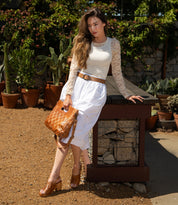 A woman in a white lace-sleeved top and skirt stands outdoors among greenery, holding the Bed Stu "Orchid" handwoven brown leather handbag with an adjustable strap.