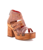 The Bed Stu Orphyx is a brown leather open-toe sandal featuring strappy details, metal studs, a thick wood platform block heel, and a back zipper closure.
