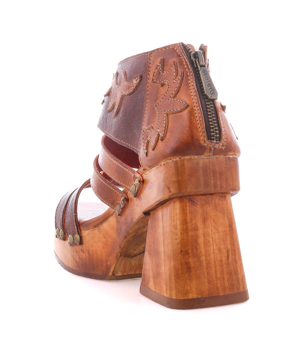 The Orphyx by Bed Stu is a brown leather high-heeled sandal with layered straps, floral stitching, metal studs, a carved wooden sole, and a back zipper, showcased here from the rear angle.