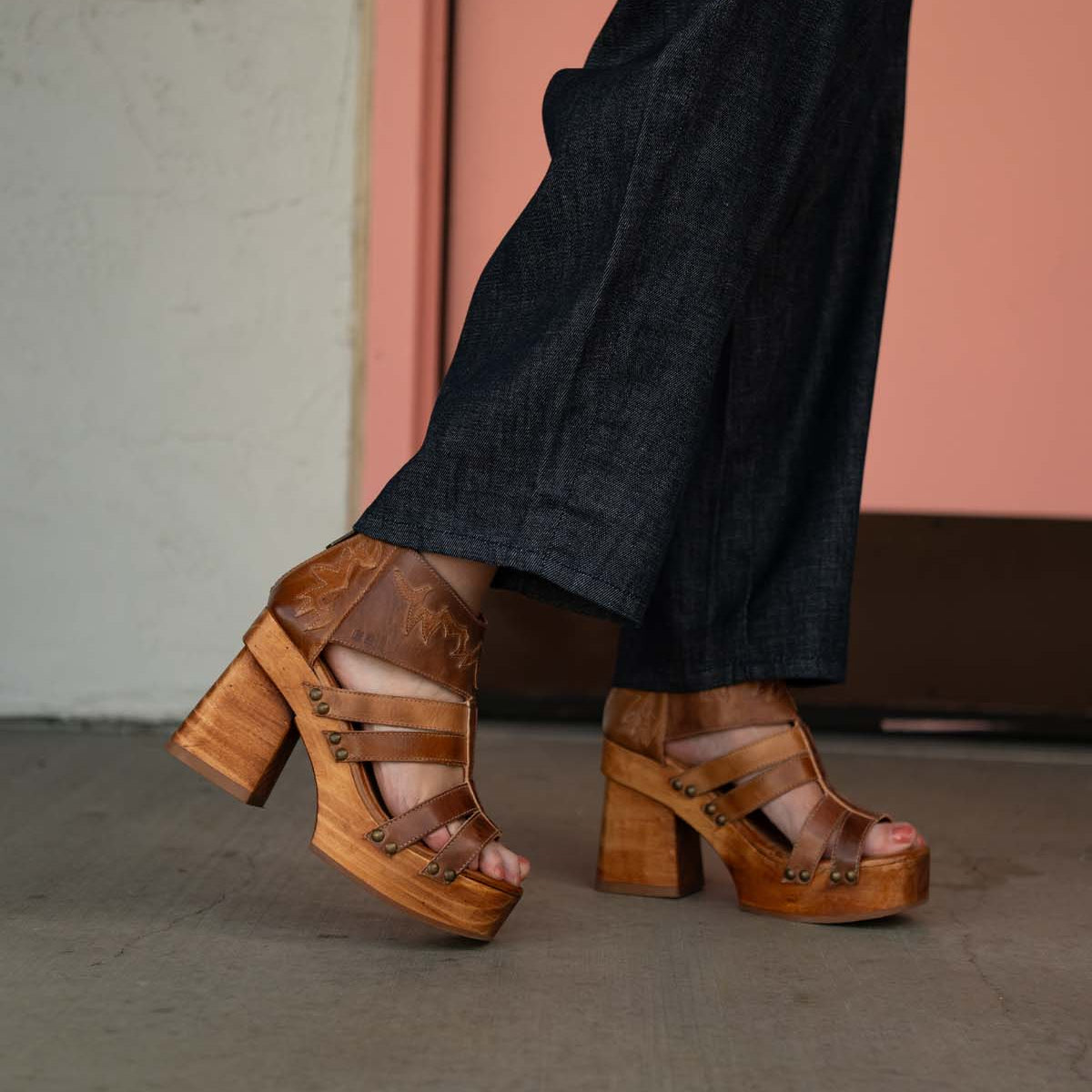 A person in dark jeans wears Bed Stu Orphyx tan platform sandals with open toes and chunky sculpted wood heels, standing on a concrete floor.