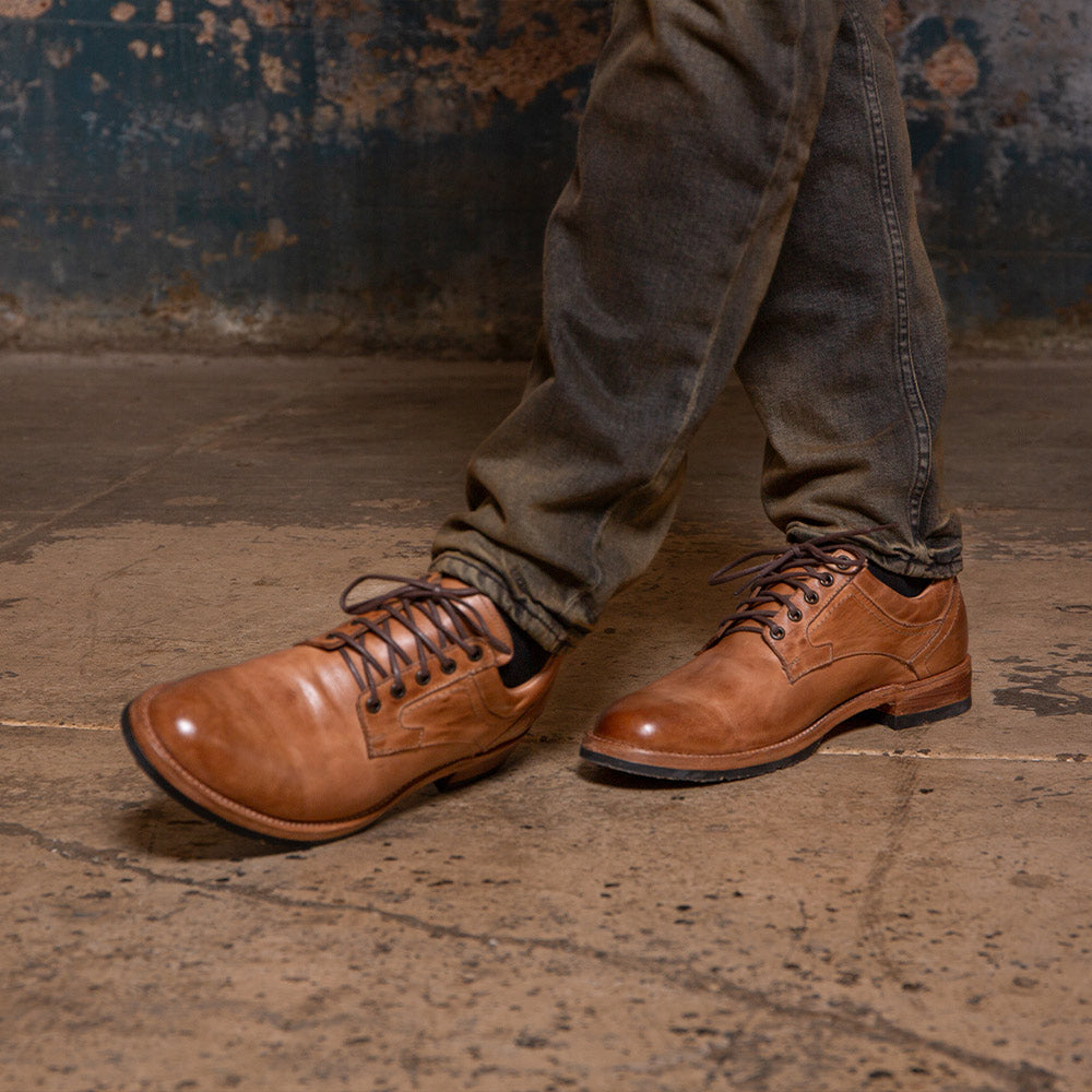 Person wearing brown leather lace-up shoes with dark pants on a concrete floor.