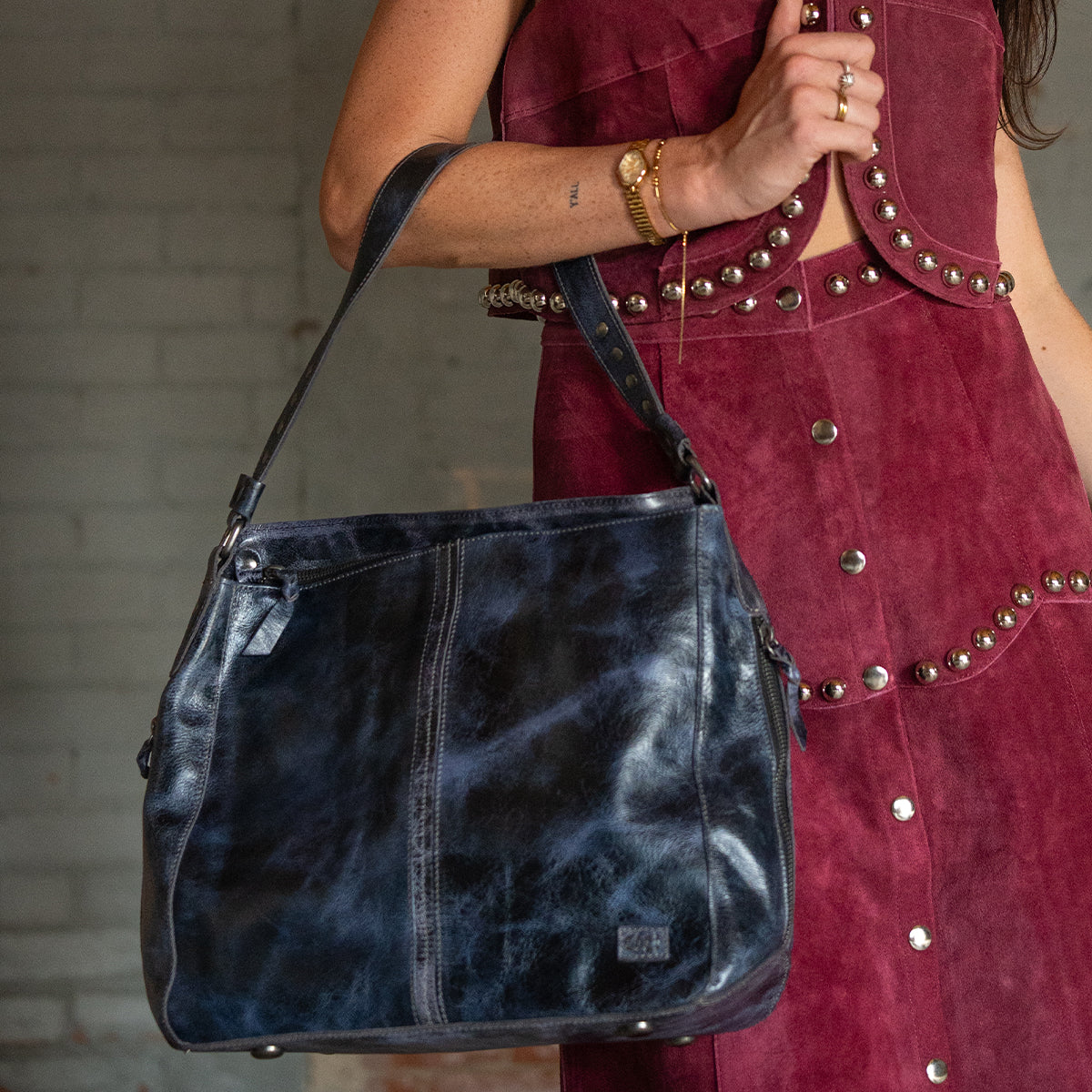 A person in a sleeveless maroon dress with metal studs holds the Bed Stu Packet LTC II large black leather shoulder tote by its strap.