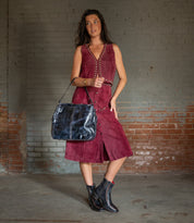 A woman in a sleeveless maroon suede dress stands indoors against a brick wall, carrying the Bed Stu Packet LTC II black leather shoulder tote and wearing black ankle boots.