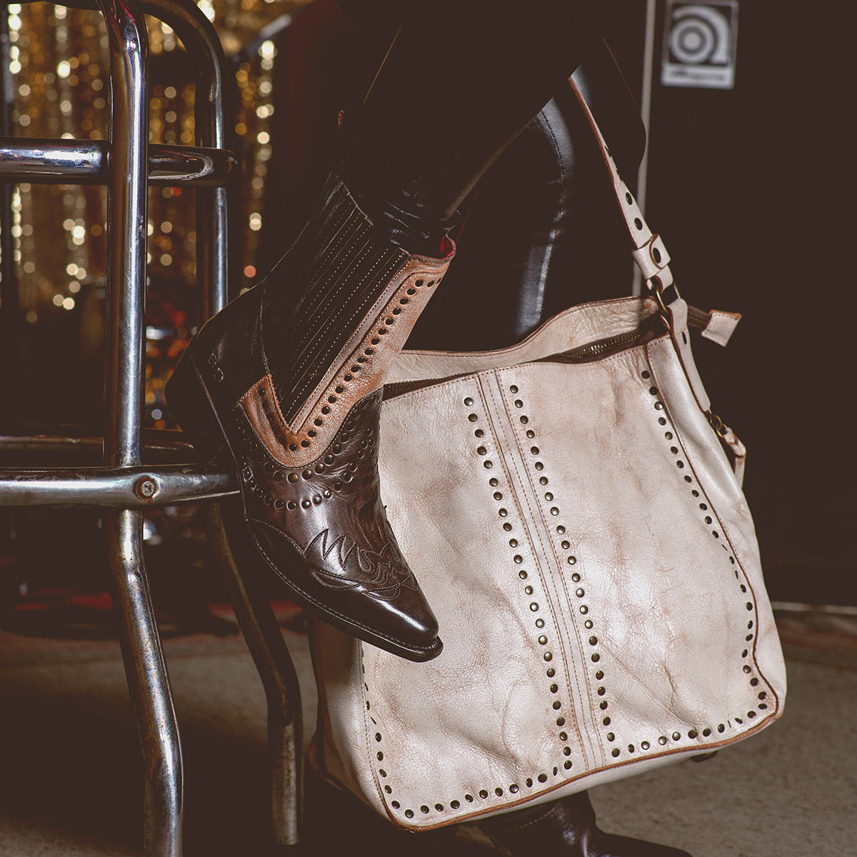 A person wearing black leather pants and brown cowboy boots sits on a stool with the Packet LTC, a large beige shoulder tote bag from Bed Stu, adorned with quality leather and metal studs resting on the floor.
