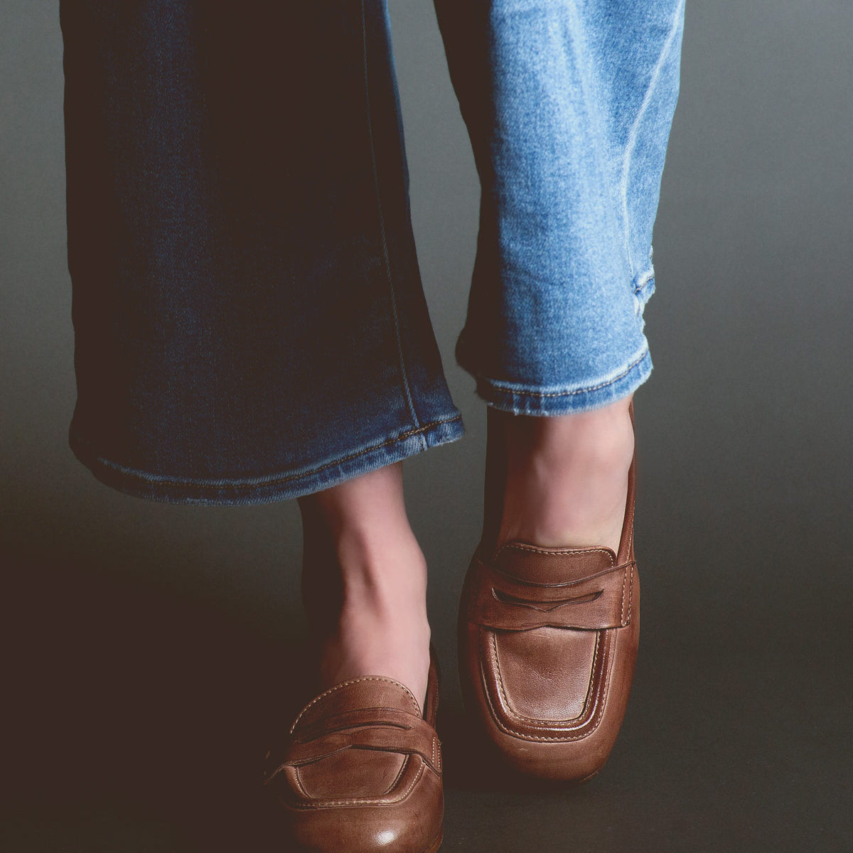 A person is wearing light blue jeans and brown Bed Stu Paris penny loafers, standing against a dark background.