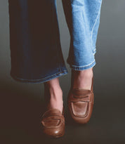 A person is wearing light blue jeans and brown Bed Stu Paris penny loafers, standing against a dark background.