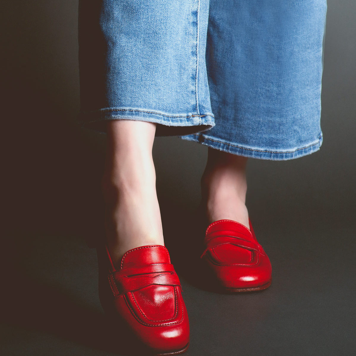 Person wearing blue jeans and red Bed Stu Paris penny loafers standing against a dark background.