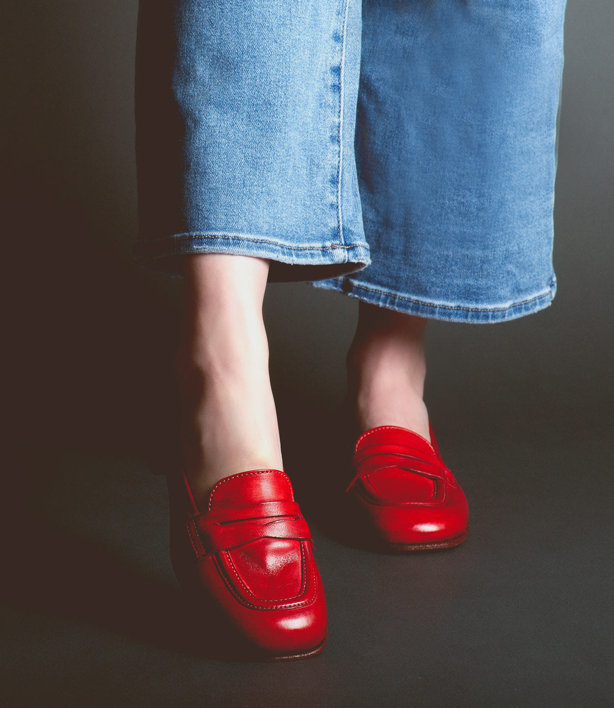 Person wearing blue jeans and red Bed Stu Paris penny loafers standing against a dark background.