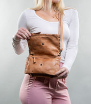 A person holds an open Bed Stu Parle brown leather shoulder bag with a crossbody strap, wearing a white top and pink pants.