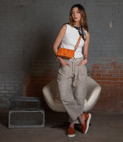 A woman in a white sleeveless top and beige pants poses stylishly against a brick wall, accessorizing with the Parle 3-in-1 orange bag from Bed Stu featuring a chic crossbody strap. Two metal cages rest artfully beside her.