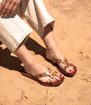 A person wearing Bed Stu Path beige leather sandals with Vibram rubber soles and cream pants sits on sandy ground.