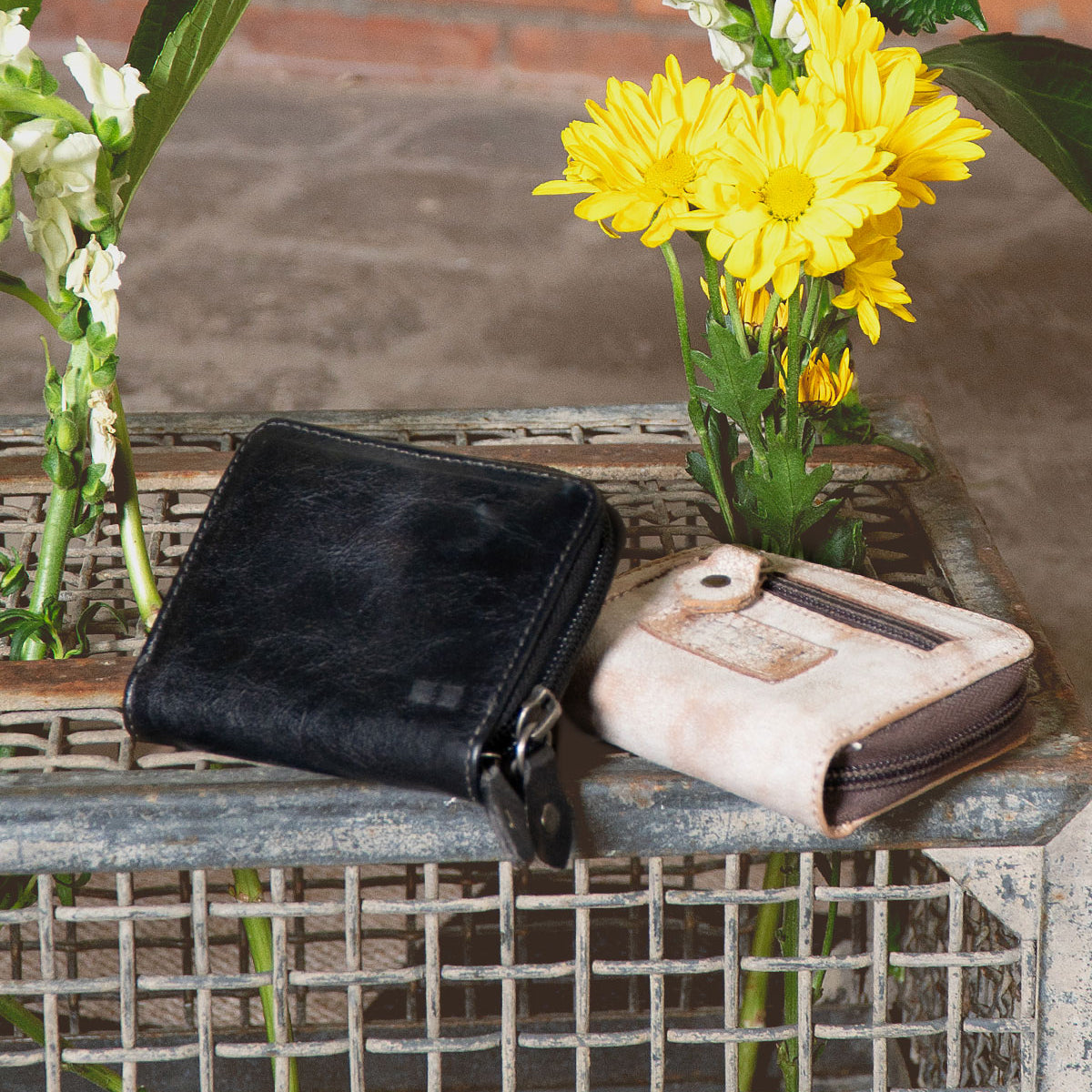 The Bed Stu Peek wallets—a black premium leather and a light brown fabric—grace a wire mesh tray, highlighting their elegant design with blooming yellow and white flowers as an elegant backdrop.