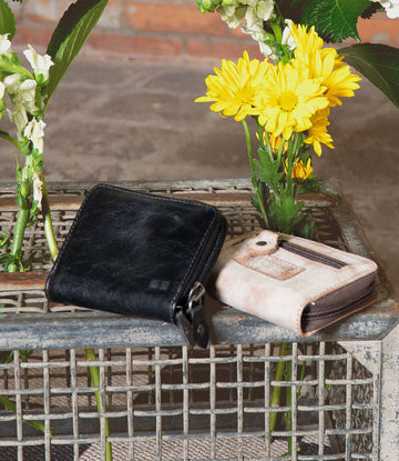 The Bed Stu Peek wallets—a black premium leather and a light brown fabric—grace a wire mesh tray, highlighting their elegant design with blooming yellow and white flowers as an elegant backdrop.