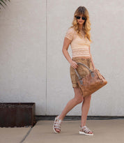 A woman stands in front of a light wall wearing sunglasses, a lace short-sleeve top, tan shorts, and Bed Stu Perfect white cushioned footbed sandals, holding a tan handbag.