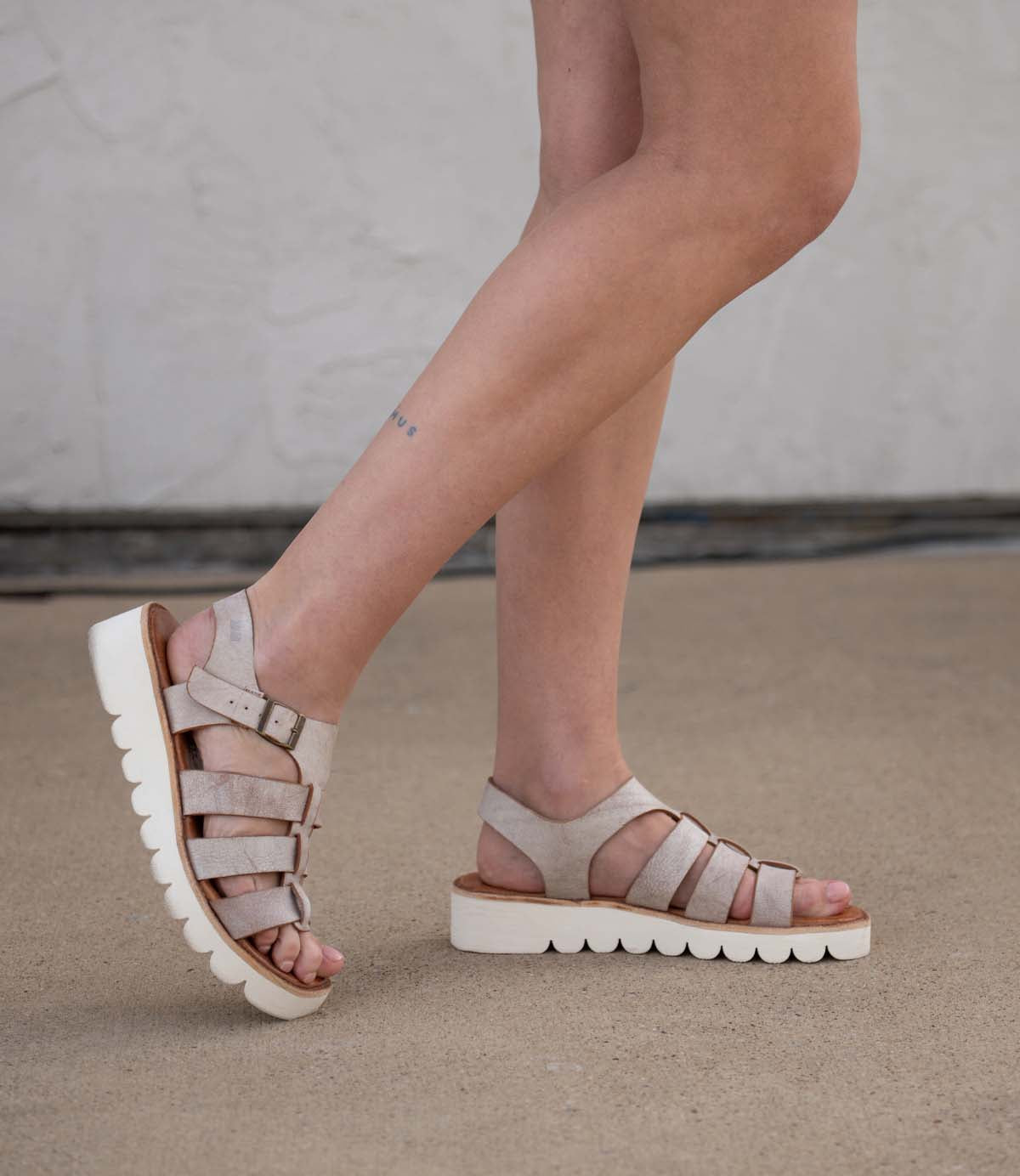 A person wears Bed Stu’s Perfect sandals in light beige, featuring thick white platform soles, while standing on a concrete surface before a light-colored wall.