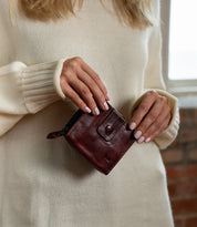 A person in a cream sweater holds the Bed|Stü Plus compact leather wallet in dark brown, featuring a zippered coin pocket and a secure snap button closure.