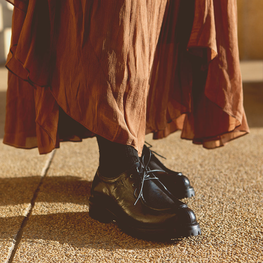 A person in a rust-colored skirt and black socks stands on a textured surface, wearing Bed Stu's handmade Portare black leather lace-up shoes, exuding a distinct 90s edge.
