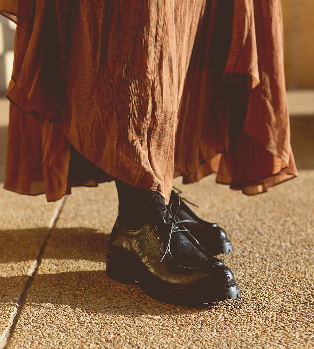 A person in a rust-colored skirt and black socks stands on a textured surface, wearing Bed Stu's handmade Portare black leather lace-up shoes, exuding a distinct 90s edge.