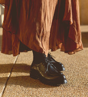 A person in a rust-colored skirt and black socks stands on a textured surface, wearing Bed Stu's handmade Portare black leather lace-up shoes, exuding a distinct 90s edge.