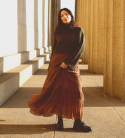 A woman in a ribbed sweater and long skirt, exuding a touch of 90s edge, stands in a sunlit corridor with stone pillars, looking towards the camera. Her Bed Stu Portare bag, crafted from vegetable-tanned leather in a handmade Italian factory, adds an air of timeless elegance to the scene.