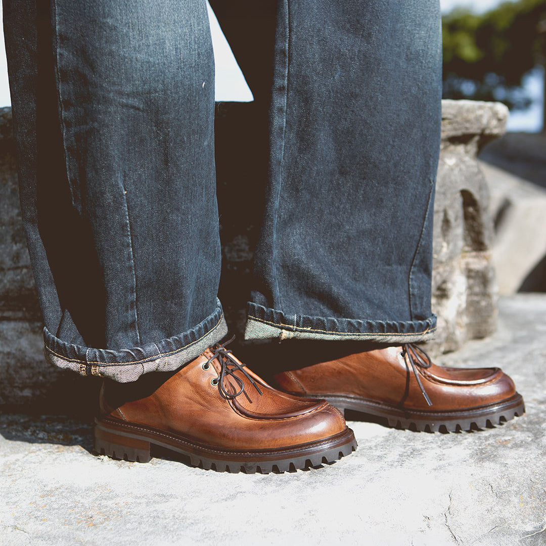 A person wearing the Portare brown leather moc-toe lace-up boots with chunky soles by Bed Stu and dark rolled-up jeans, standing on a stone surface, exudes Italian artistry and vintage-inspired style.
