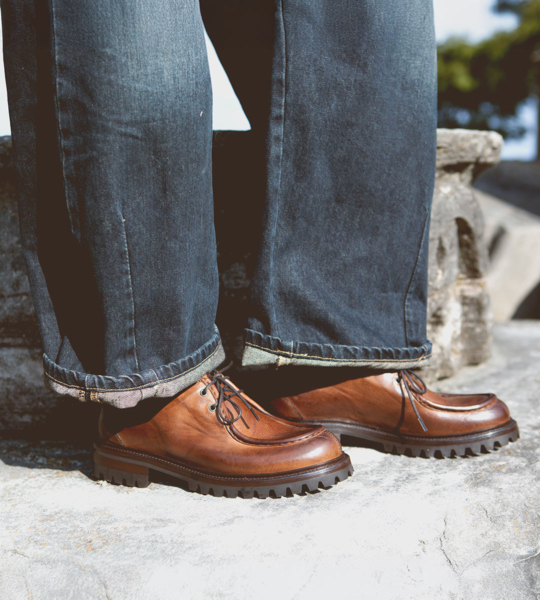 A person wearing the Portare brown leather moc-toe lace-up boots with chunky soles by Bed Stu and dark rolled-up jeans, standing on a stone surface, exudes Italian artistry and vintage-inspired style.