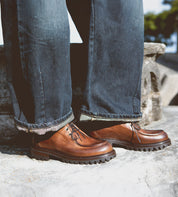 A person wearing the Portare brown leather moc-toe lace-up boots with chunky soles by Bed Stu and dark rolled-up jeans, standing on a stone surface, exudes Italian artistry and vintage-inspired style.