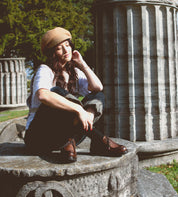 A woman wearing the Portare hat from Bed Stu, crafted from beige vegetable-tanned leather, sits on a stone structure amid columns and lush greenery under the sunlight. This scene exudes timeless elegance and craftsmanship, echoing a handmade Italian factory with a hint of 90s flair.