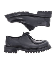 Two Portare shoes by Bed Stu in black leather, featuring thick, rugged soles, are presented vertically with the soles touching against a white background. These vintage-inspired pieces, crafted in an Italian factory, infuse a 90s edge with timeless style and modern durability.