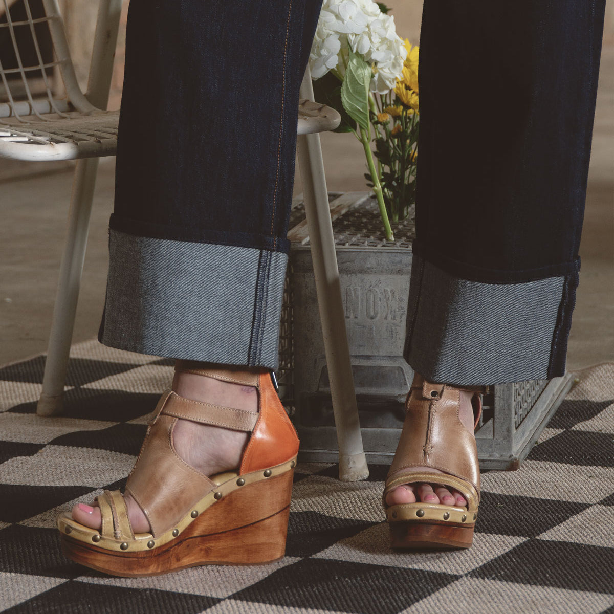 Someone wearing Bed Stu's Princess tan wedge sandals with strappy uppers and dark rolled-up jeans stands on a checkered rug. A metal container filled with flowers adds a touch of nature to this stylish scene.