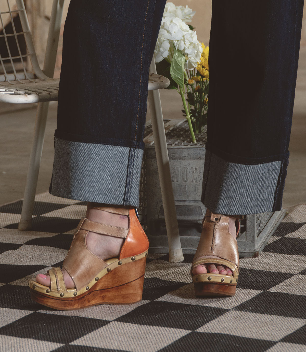 Someone wearing Bed Stu's Princess tan wedge sandals with strappy uppers and dark rolled-up jeans stands on a checkered rug. A metal container filled with flowers adds a touch of nature to this stylish scene.