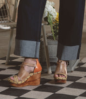 Someone wearing Bed Stu's Princess tan wedge sandals with strappy uppers and dark rolled-up jeans stands on a checkered rug. A metal container filled with flowers adds a touch of nature to this stylish scene.