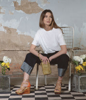 A woman elegantly sits on a chair, clutch in hand, surrounded by vibrant flowers. She wears a white t-shirt, dark pants, and Bed Stu's Princess sandals featuring woven strappy uppers. The scene is completed by the rustic charm of a distressed wall in the background.